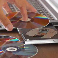 Ręka wkłada płytę do napędu laptopa. Obok leżą kolejne płyty, gotowe do nagrania za pomocą programu do nagrywania płyt.