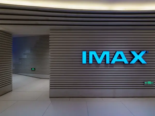 Jasnoniebieski napis IMAX na szarej, prążkowanej ścianie. To wejście do kina, gdzie czeka Cię niezapomniane wrażenie.